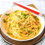 Noodles in a bowl topped with green onions and sesame seeds with red chopsticks across the side of the bowl.