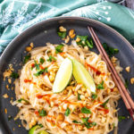 Noodles in a bowl topped with cilantro, sriracha, and lime wedges with chopsticks.