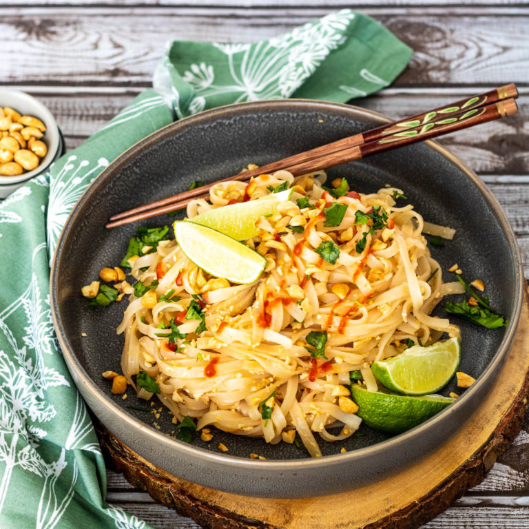 Noodles in a bowl topped with cilantro, sriracha, and lime wedges with chopsticks.