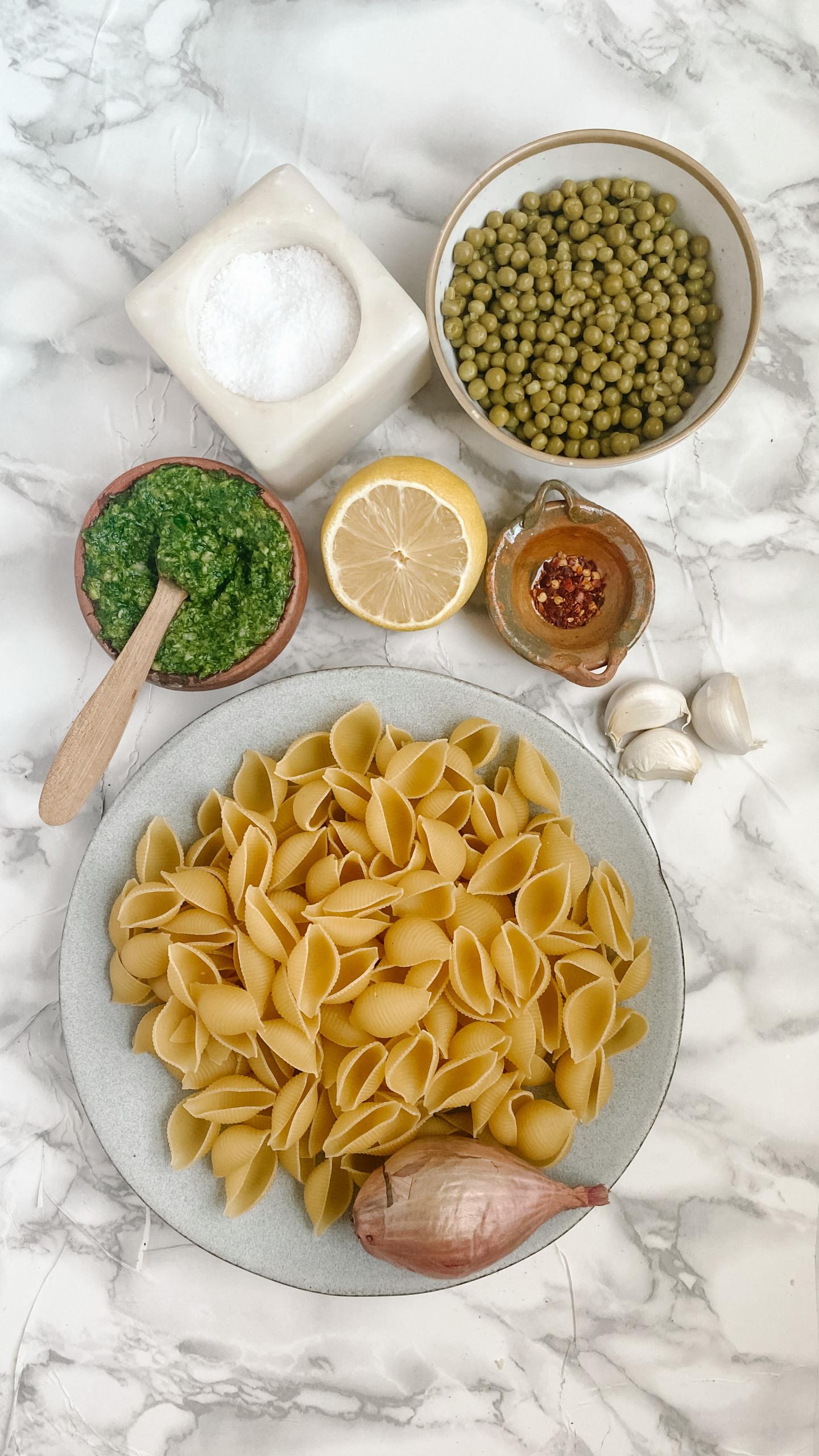 Bowls with pasta shells, peas, pesto, a lemon, red pepper flakes, a shallot, salt, and garlic on a white marble counter.