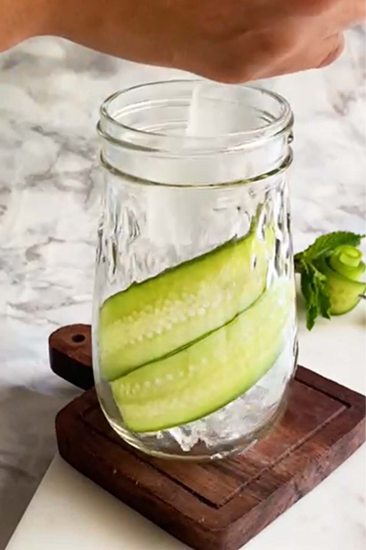 Ice is dropped into a glass with cucumber ribbons along the side.