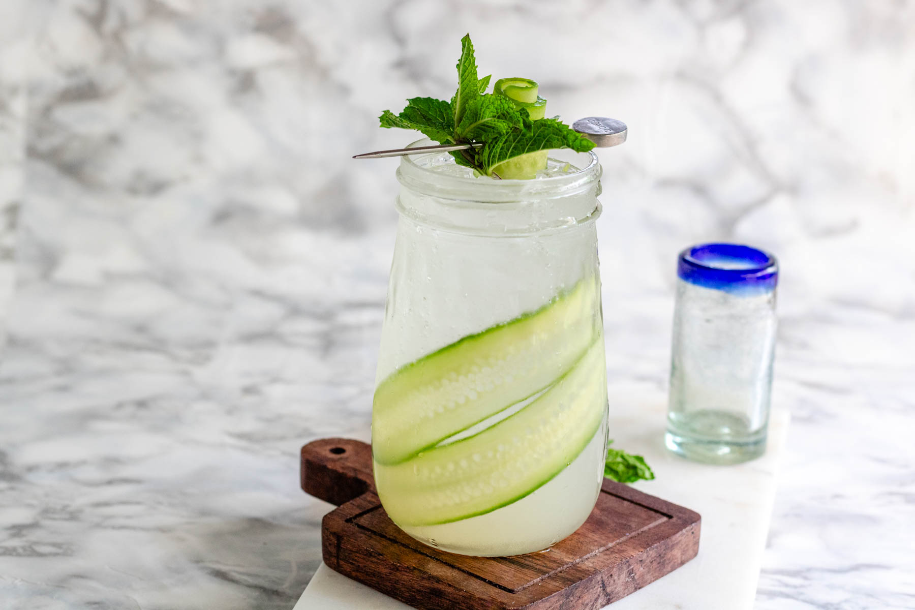 A tapered glass holds a clear drink with cucumber ribbons in it and a mint cucumber garnish on top.