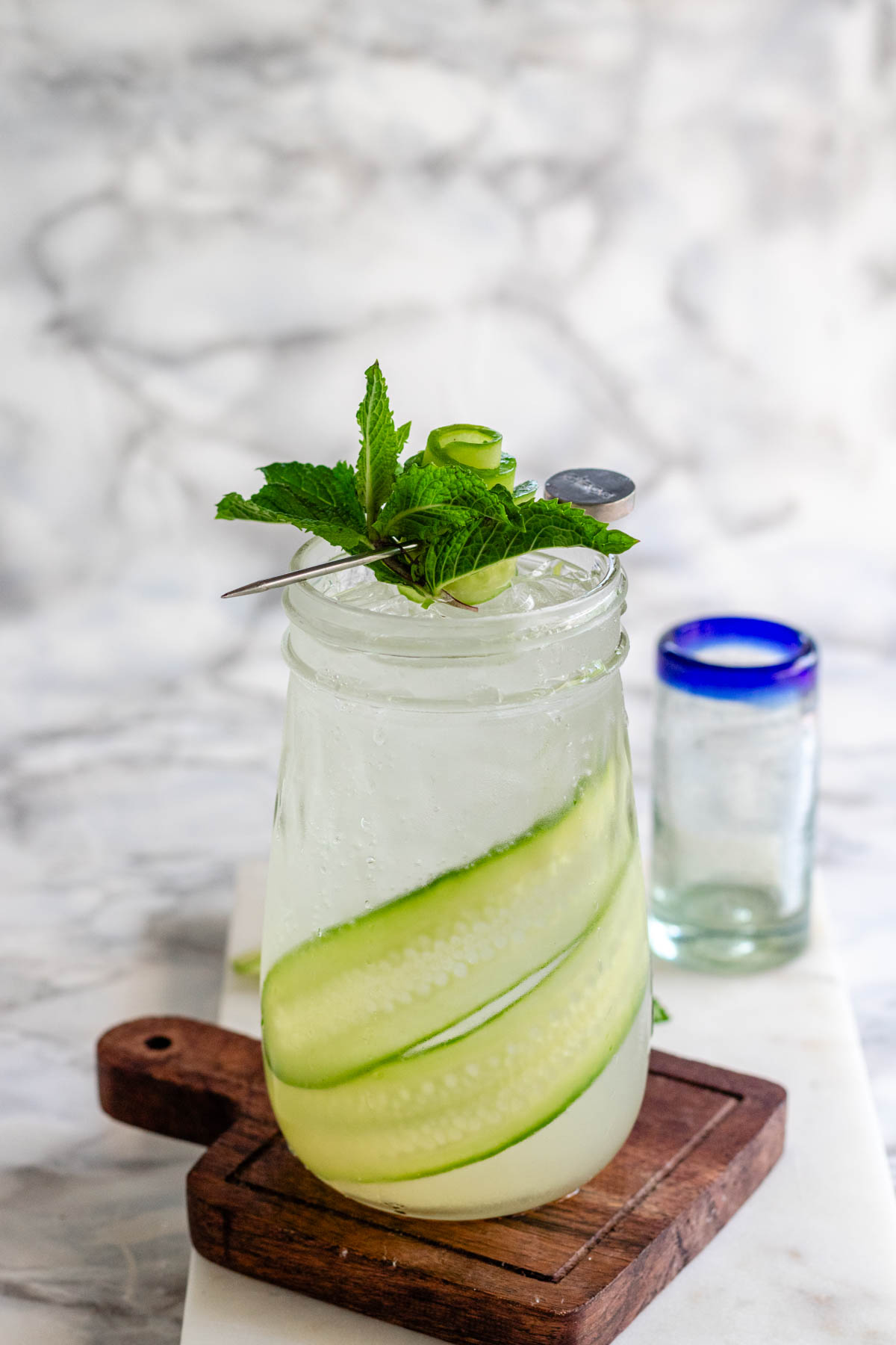 A tapered glass holds a clear drink with cucumber ribbons in it and a mint cucumber garnish on top.