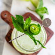 A tapered glass holds a clear drink with cucumber ribbons in it and a mint cucumber garnish on top with a text title for Pinterest.