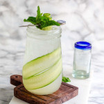 A tapered glass holds a clear drink with cucumber ribbons in it and a mint cucumber garnish on top.