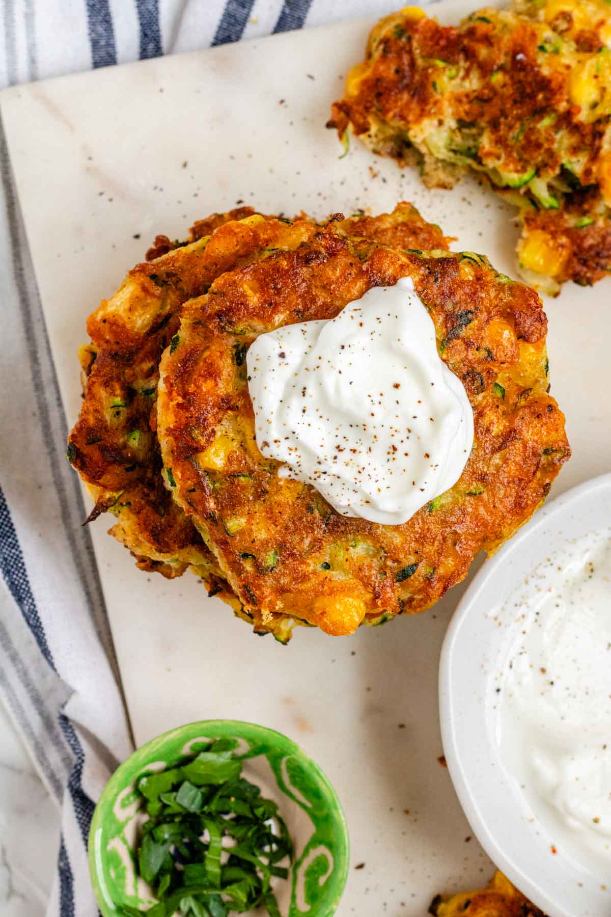 Golden brown zucchini corn fritters stacked on a marble tray with sour cream.