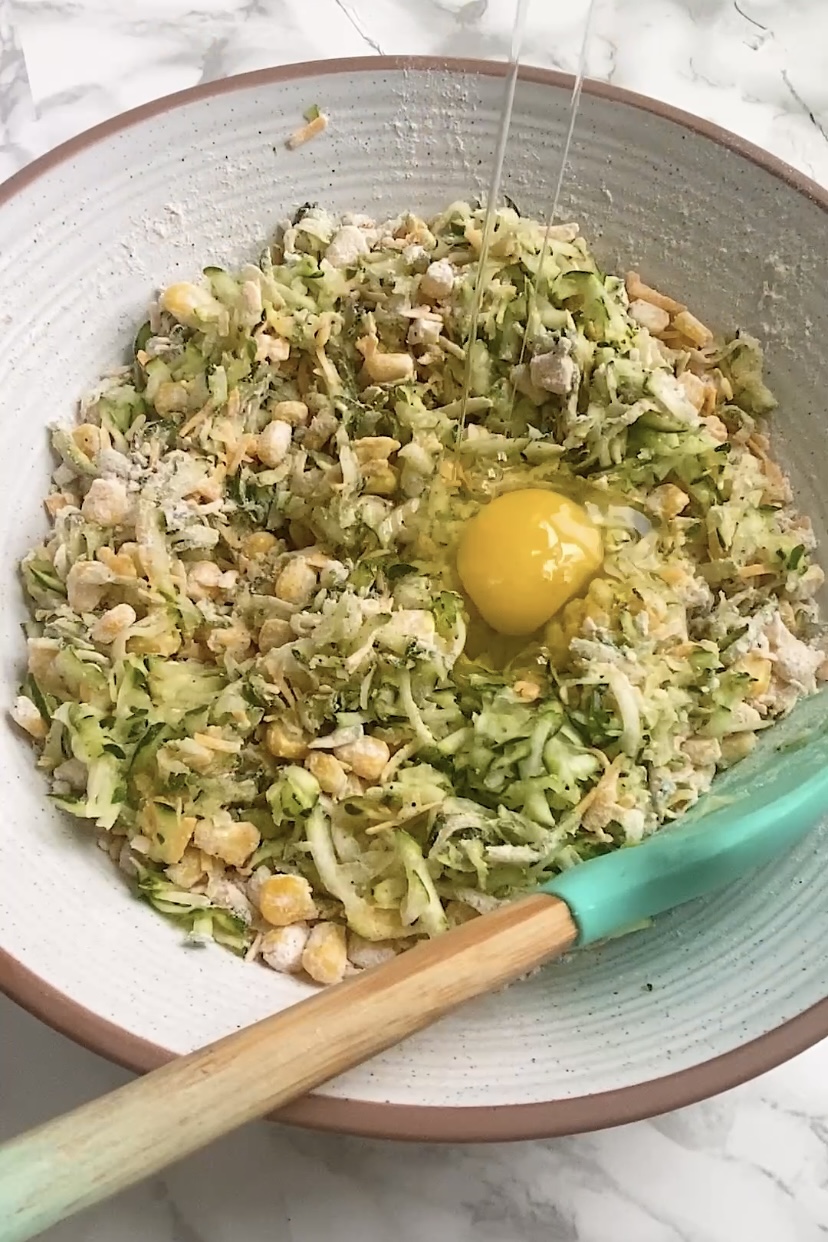 An egg is added to a large bowl with corn, flour, and zucchini in it.