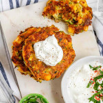 Golden brown zucchini corn fritters stacked on a marble tray with sour cream.