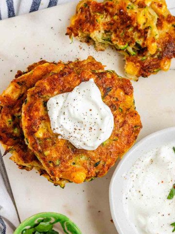 Golden brown zucchini corn fritters stacked on a marble tray with sour cream.