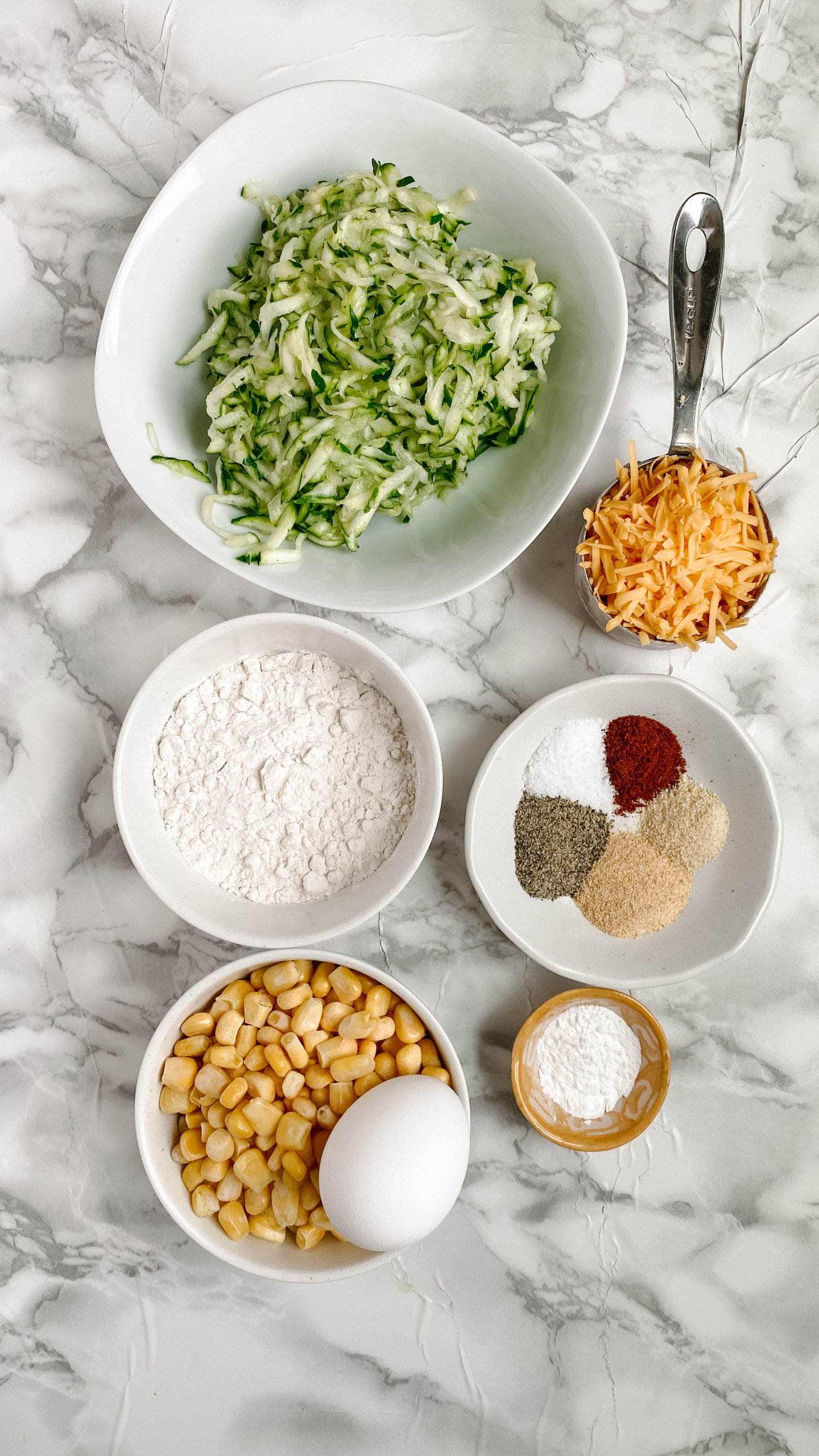 Ingredients in bowls for zucchini and corn fritters on a white marble surface.