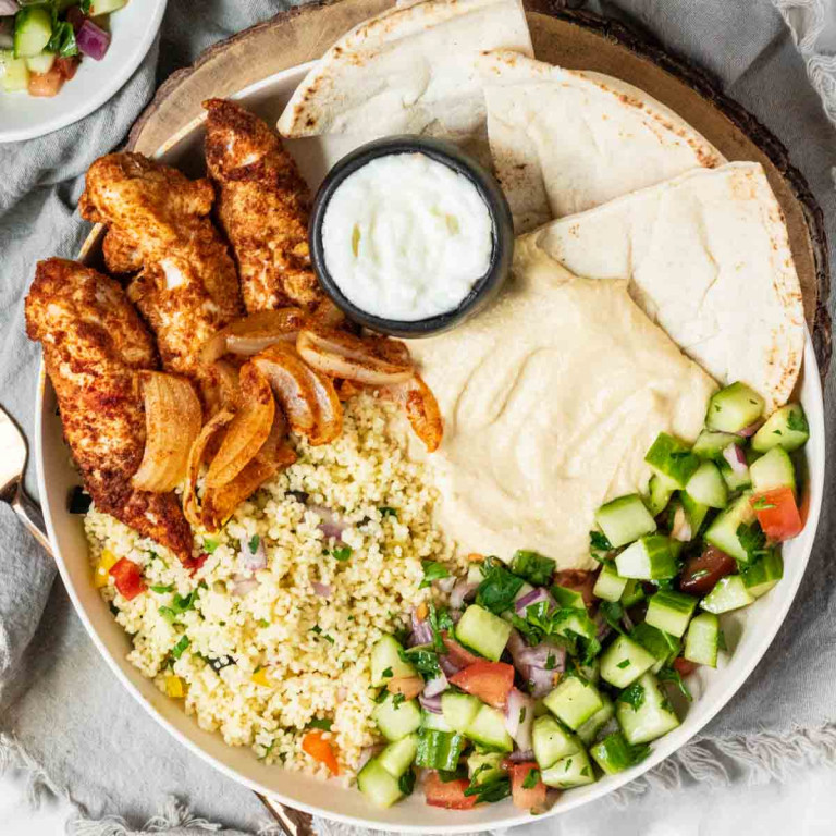 A chicken shawarma plate with chicken, pita, sauces, couscous and cucumber salad.