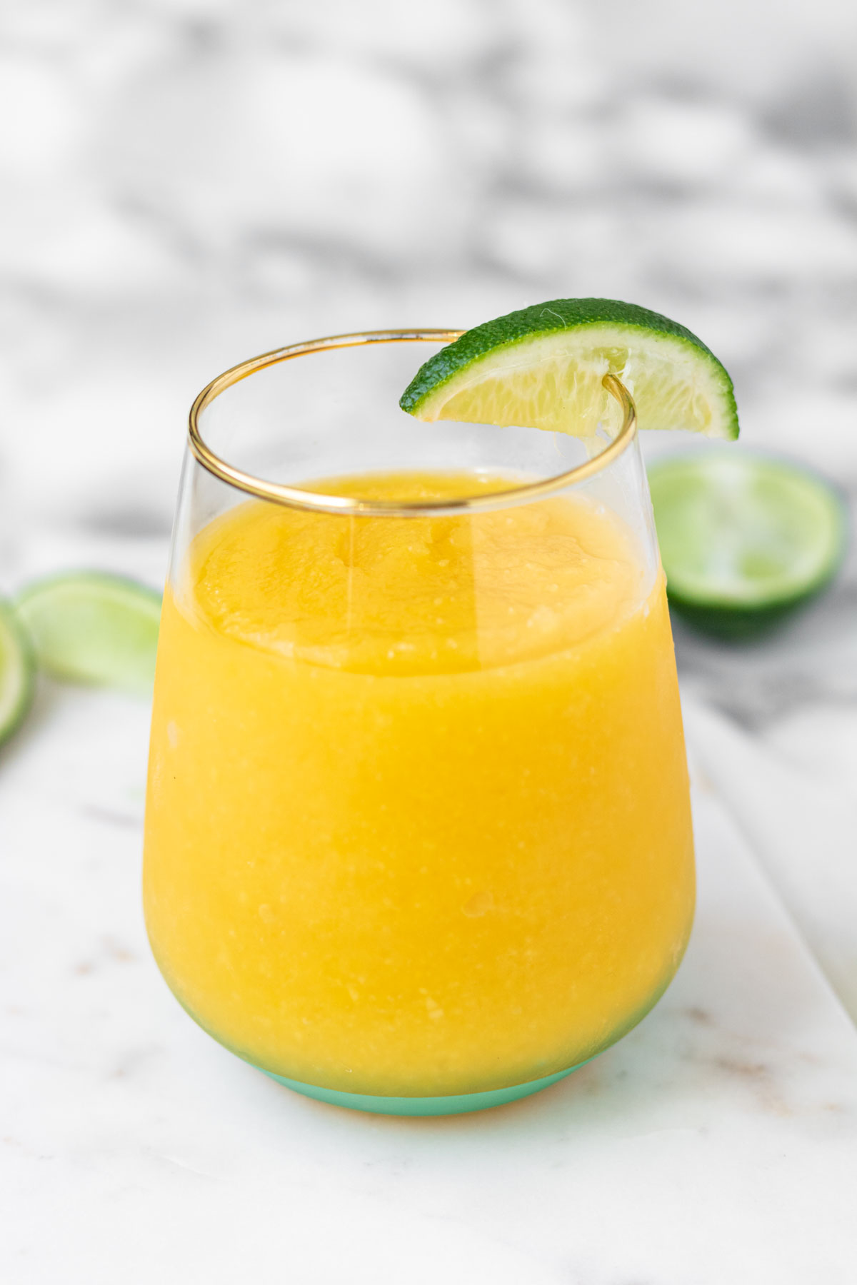A frozen mango margarita in a glass on a marble background with lime wedges.
