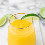 A frozen mango margarita in a glass on a marble background with lime wedges.