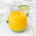 A frozen mango margarita in a glass on a marble background with lime wedges.