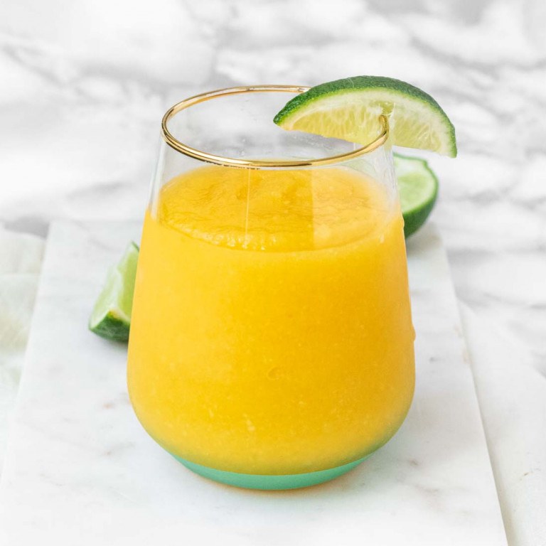 A frozen mango margarita in a glass on a marble background with lime wedges.