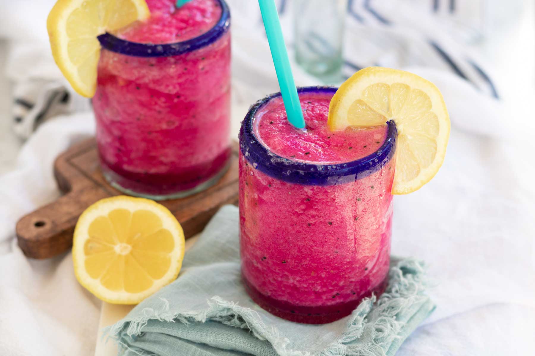 A bright pink blended margarita in a blue-rimmed glass with a blue straw and lemon garnish.