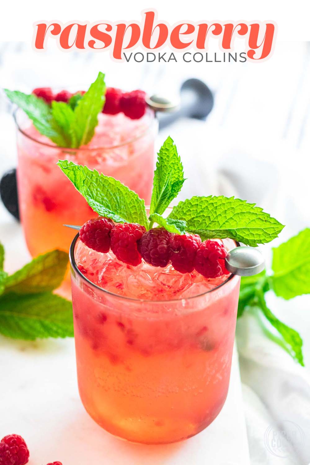 A pink drink in a pink glass garnished with raspberries on a cocktail pick and fresh mint.