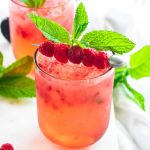 A pink drink in a pink glass garnished with raspberries on a cocktail pick and fresh mint.