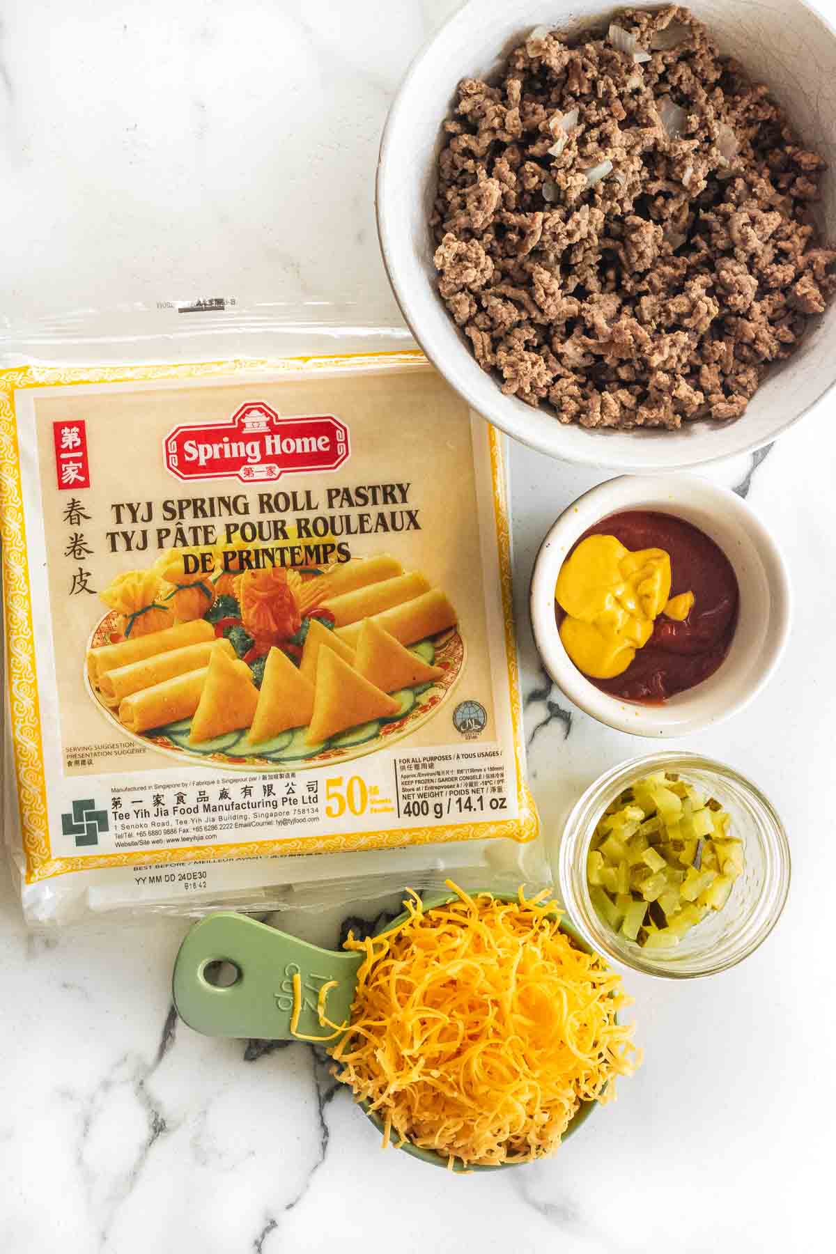 Ingredients to make cheeseburger spring rolls on a white marble counter.