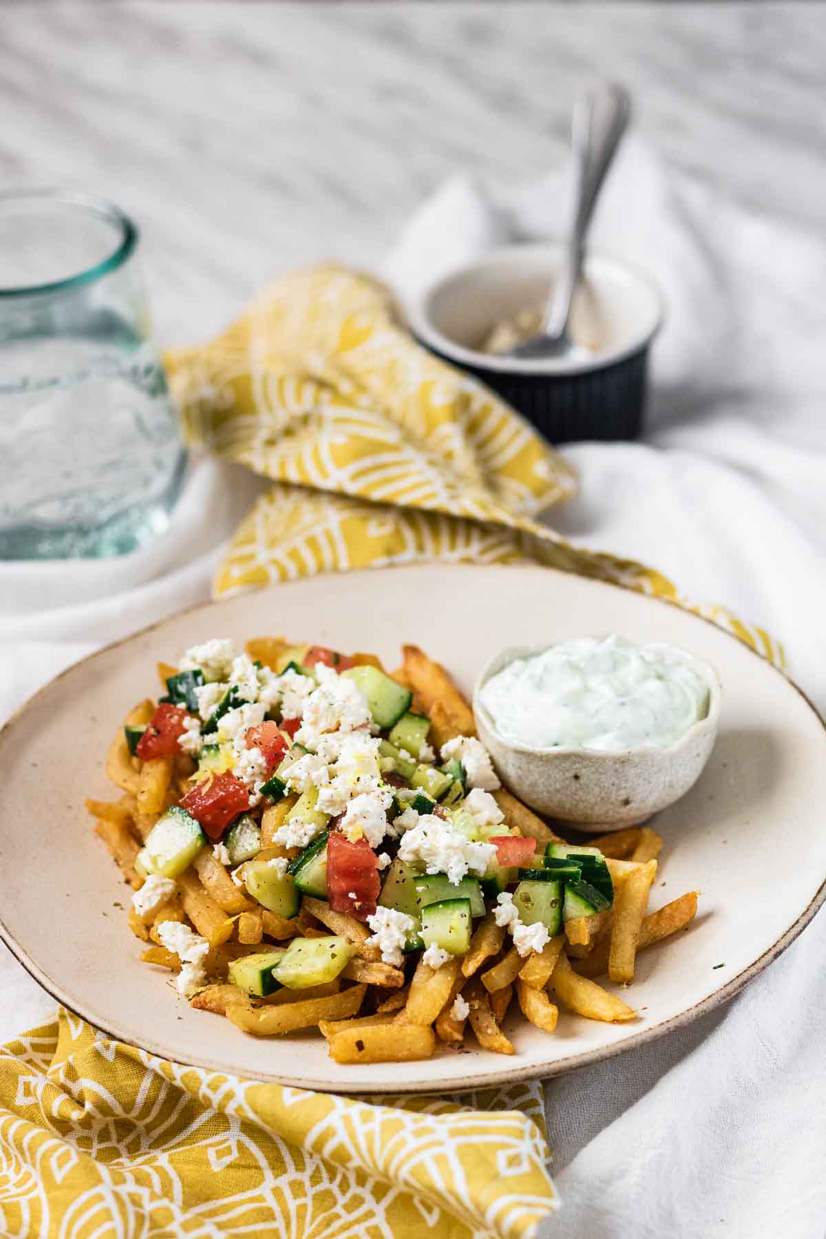 Fries on a plate topped with cucumber, tomato and feta cheese with a bowl of Tzatziki sauce.