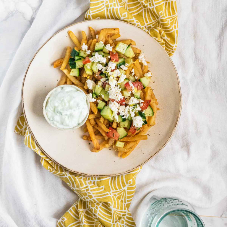Fries on a plate topped with cucumber, tomato and feta cheese with a bowl of Tzatziki sauce.
