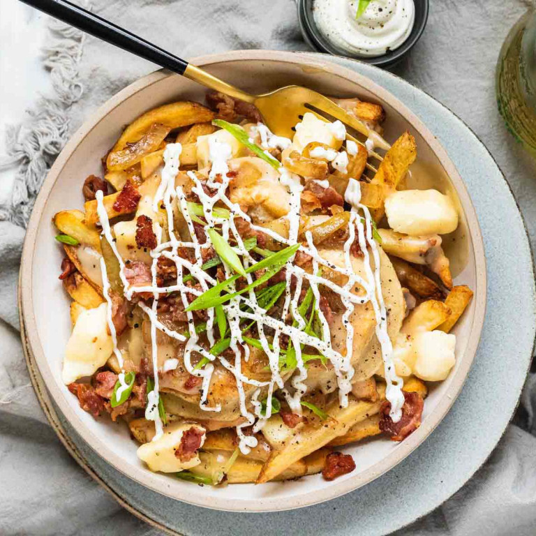 Fries, perogies, cheese curds, bacon, green onions, and drizzled sour cream in a bowl.