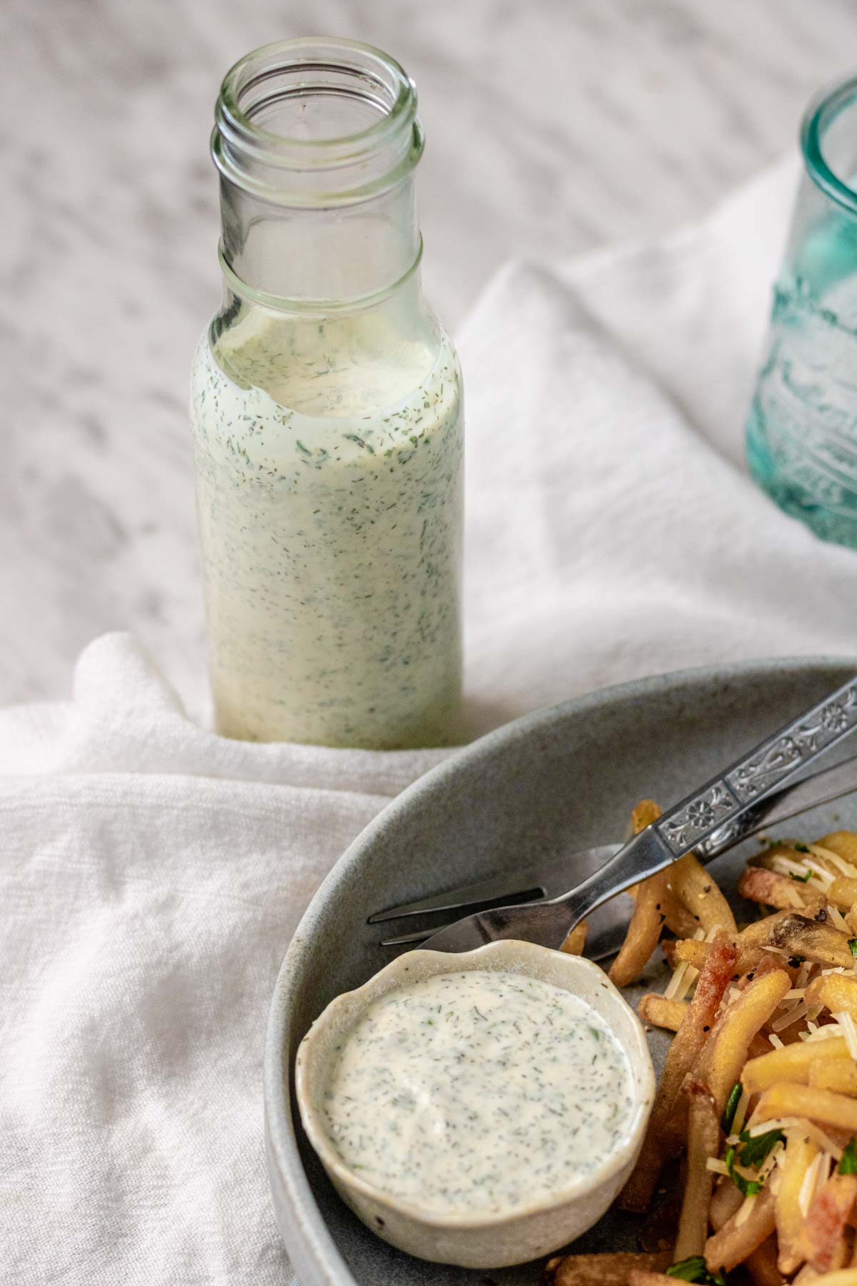 Homemade ranch dressing in a tall glass bottle with fries and a bowl of dip in the foreground.