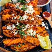 Baked sweet potato wedges on a black slate platter, covered in feta, fresh oregano, and a drizzle of red sauce.