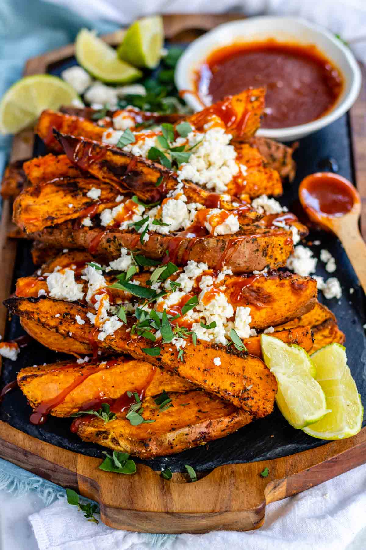 Baked sweet potato wedges on a black slate platter, covered in feta, fresh oregano, and a drizzle of red sauce.