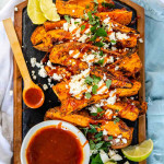 Baked sweet potato wedges on a black slate platter, covered in feta, fresh oregano, and a drizzle of red sauce.