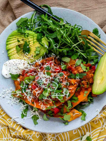 A plate with a sweet potato topped with enchilada sauce and vegetables with avocado and greens on the side.