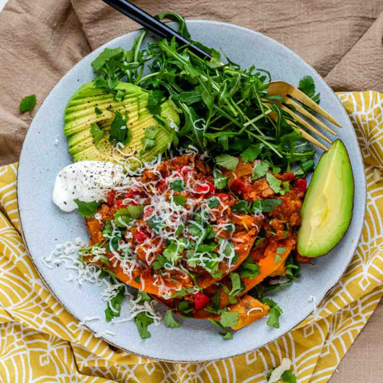 A plate with a sweet potato topped with enchilada sauce and vegetables with avocado and greens on the side.