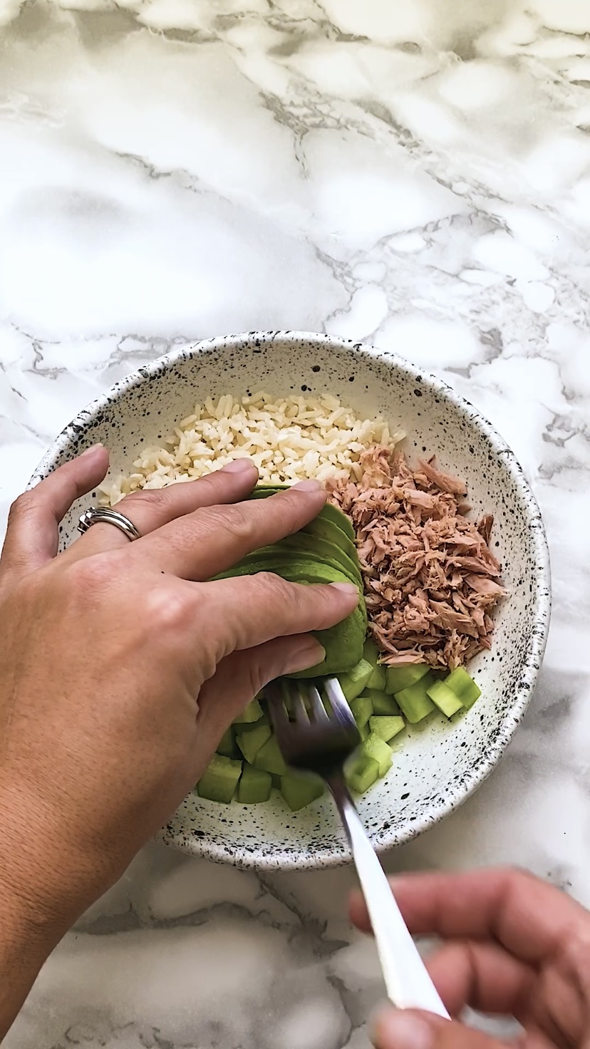 Adding avocado to a bowl of tuna, rice, and cucumber.