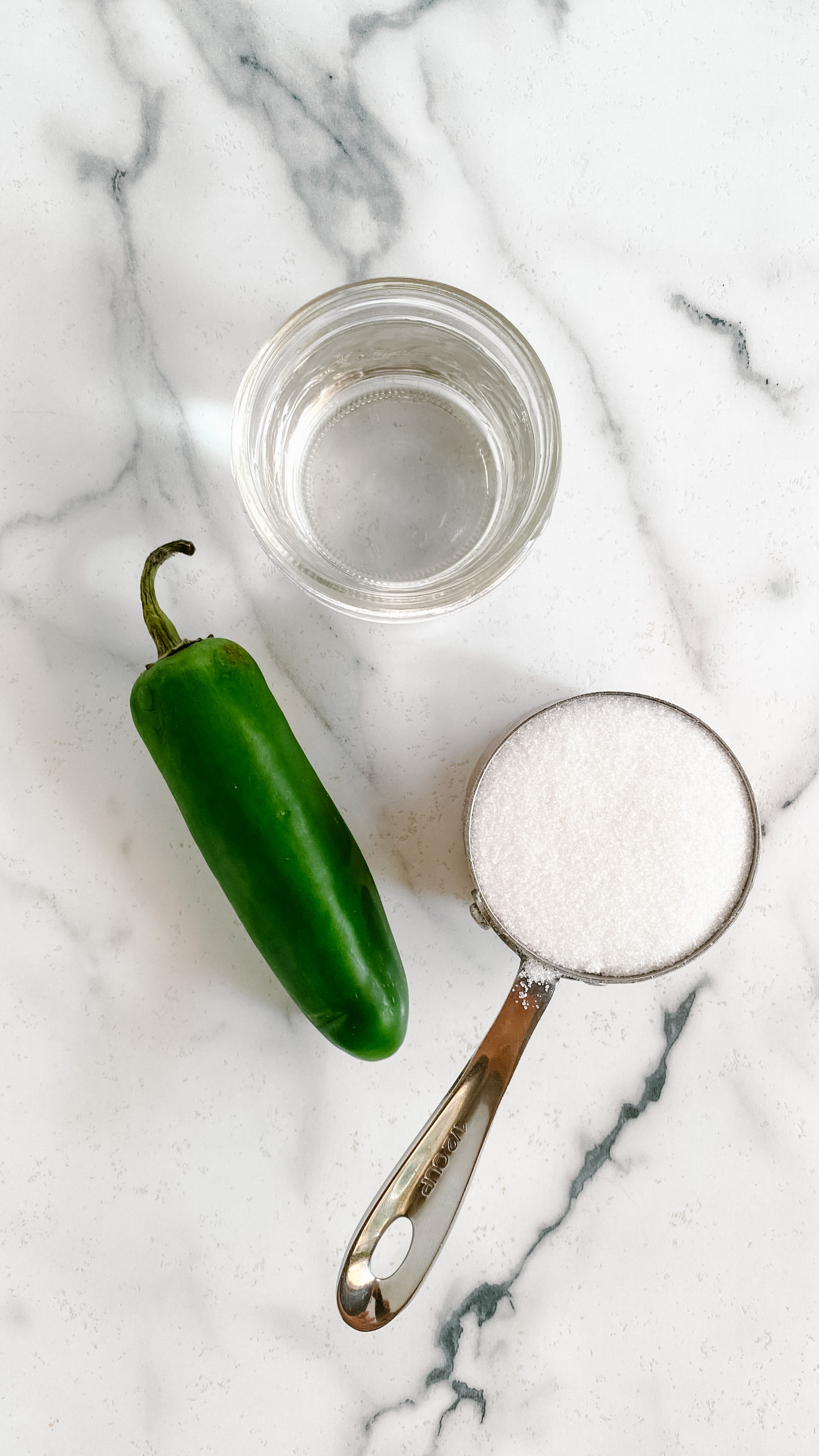 A jar of water, a measuring cup of sugar, and a jalapeno on a white marble background.