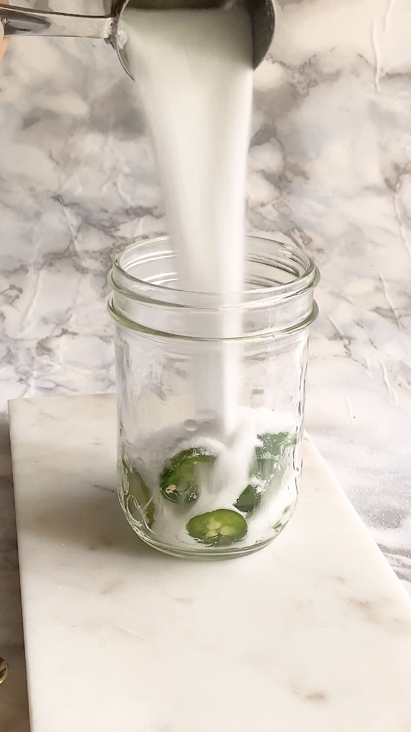 Sugar is poured into a jar with jalapeno slices in it.