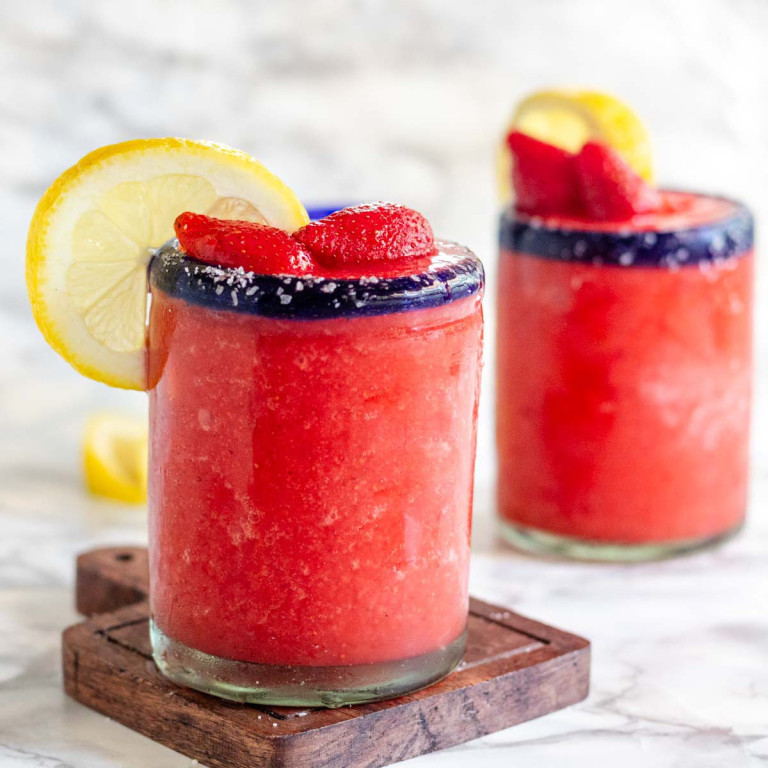 A frozen pink drink in a glass garnished with strawberries and a lemon slice.