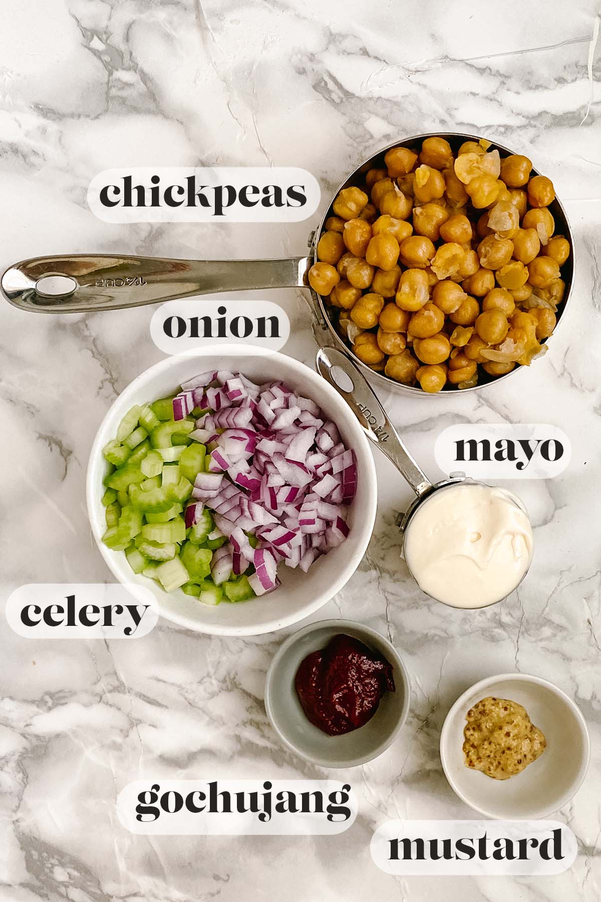 Ingredients for a chickpea salad sandwich spread sit on a white marble counter with text labels.