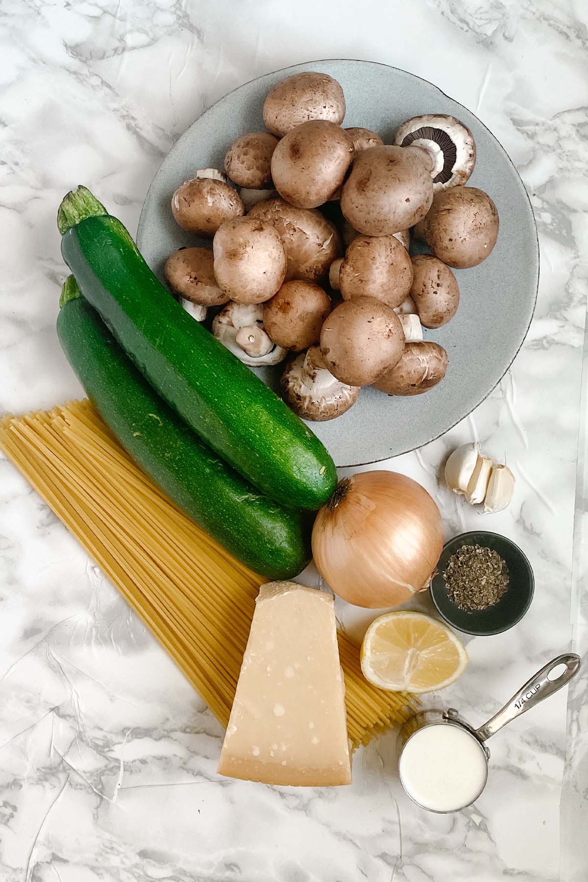 Ingredients for mushroom and zucchini pasta on a white marble surface.