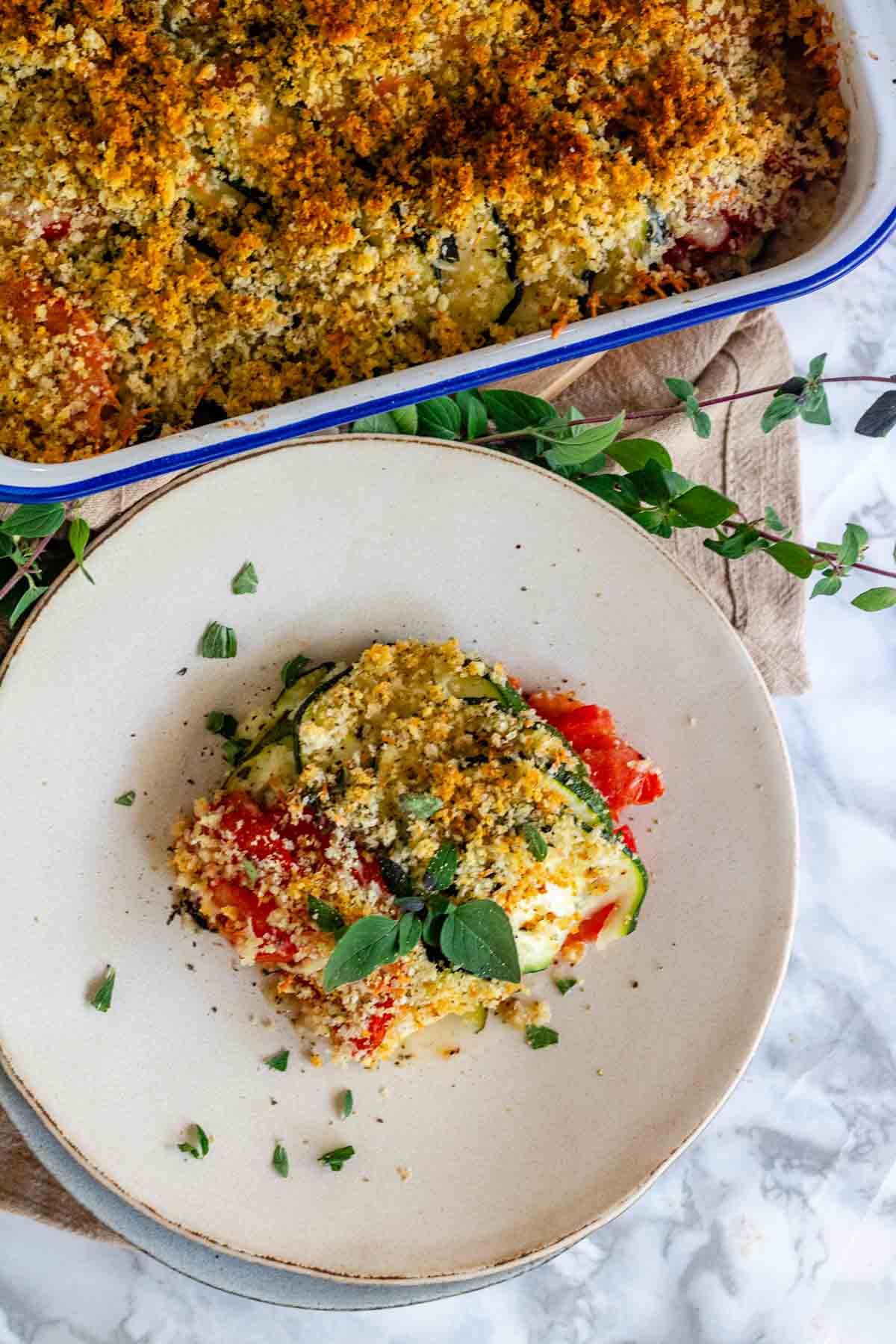 A baked casserole on a plate topped with browned bread crumbs and oregano leaves.
