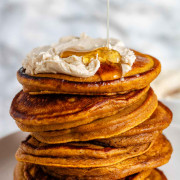 Maple syrup is poured onto a stack of pumpkin pancakes topped with whipped butter.