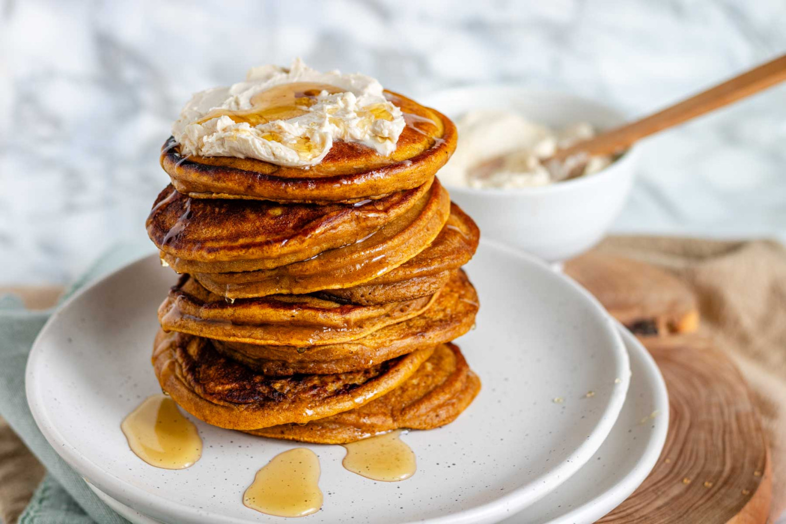 A tall stack of golden-brown pancakes topped with cream and drizzled with syrup, served on a speckled white plate.