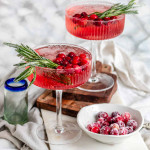 A pink cocktail in glasses garnished with sugared cranberries and rosemary.
