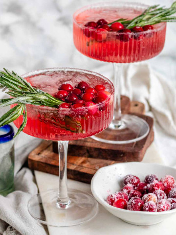 A pink cocktail in glasses garnished with sugared cranberries and rosemary.