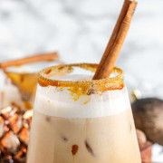 A creamy drink in a rimmed glass with a cinnamon stick in it with text for Pinterest.