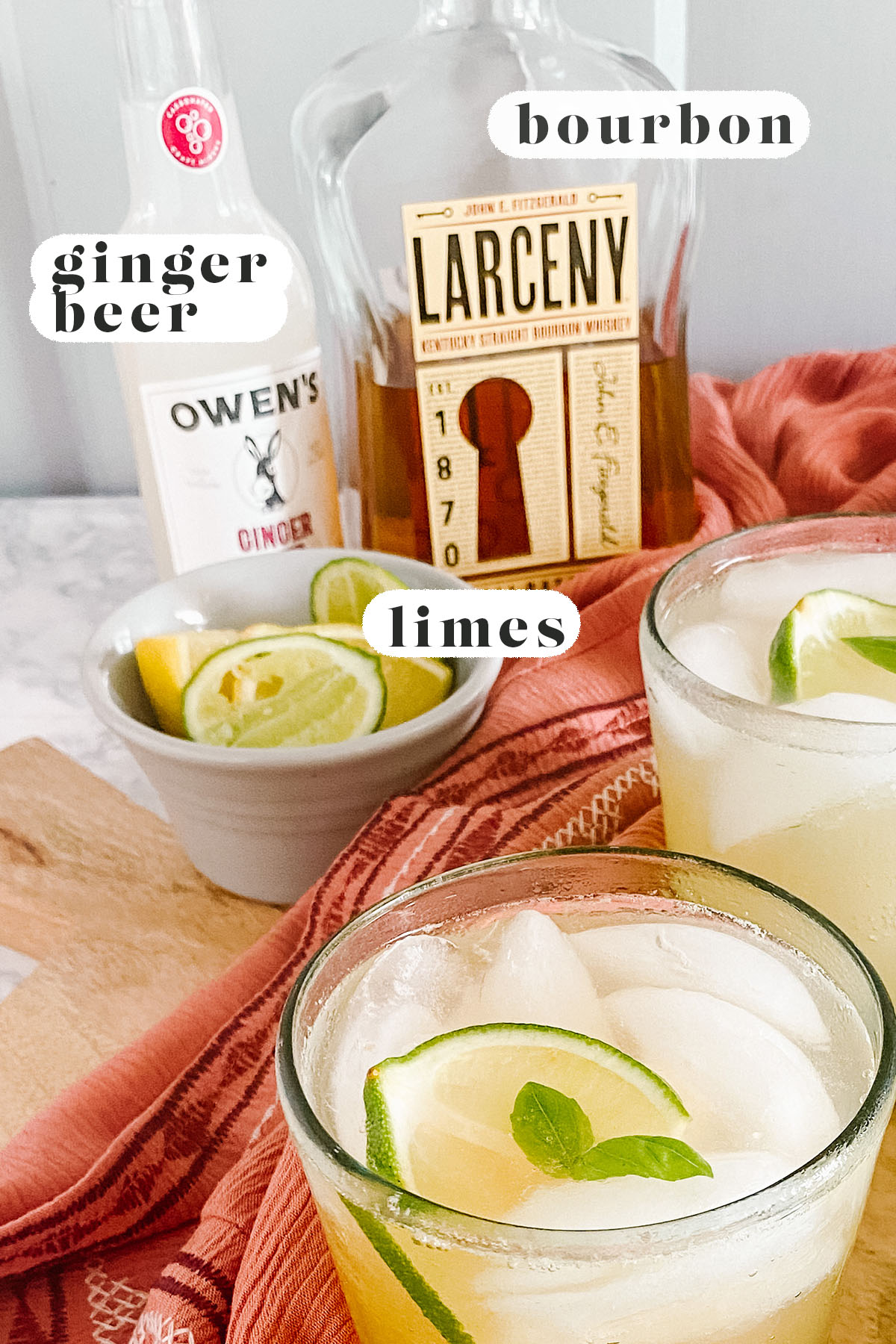 A cocktail in a glass garnished with lime and mint with lime, ginger beer, and bourbon in the background.