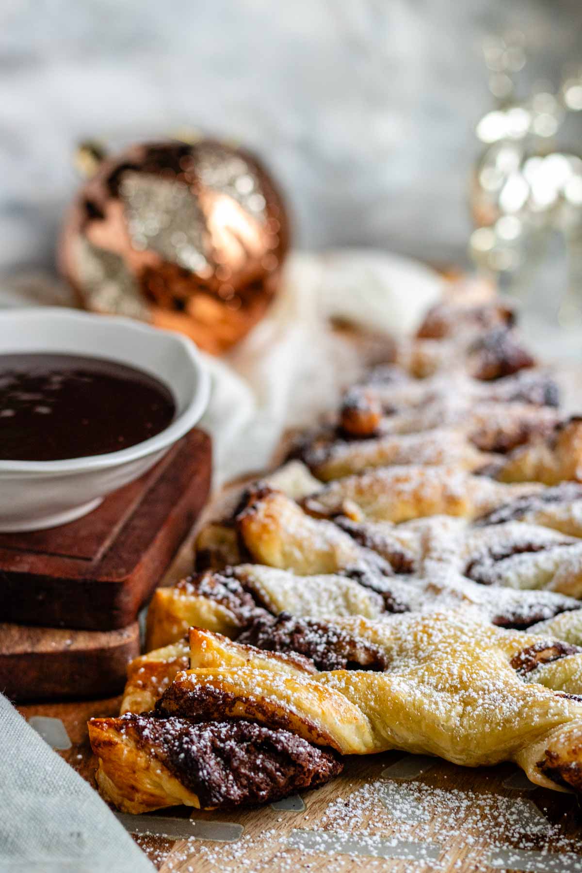 A Nutella puff pastry tree dusted with icing sugar on a wooden board with a bowl of chocolate dip.