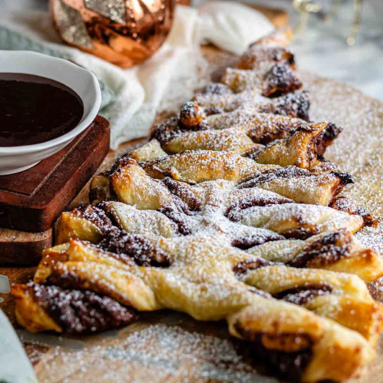A Nutella puff pastry tree dusted with icing sugar on a wooden board with a bowl of chocolate dip.