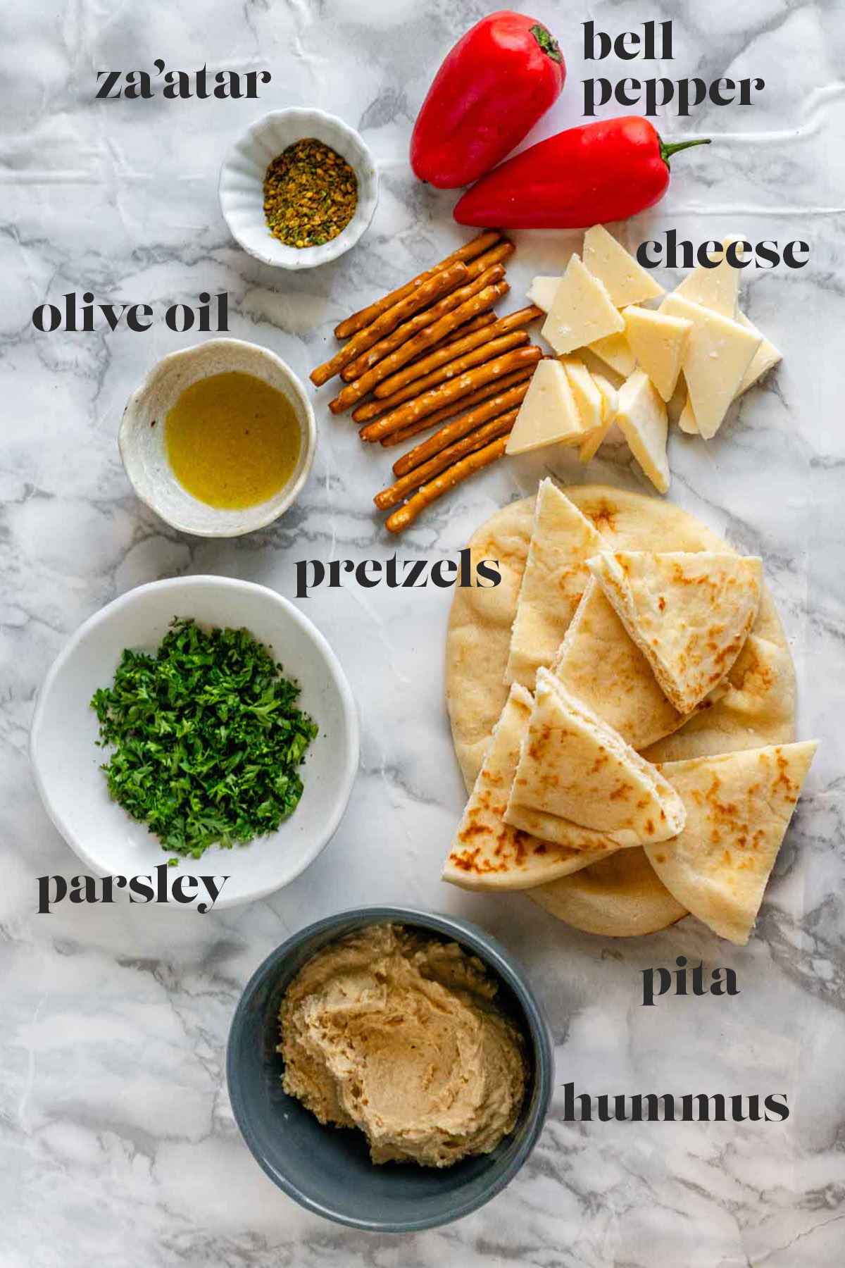Cut pitas, a bowl or hummus, a bowl of parsley, a bowl or oil, a bowl of za'atar, bell peppers, pretzels, and cheese on a white marble counter.