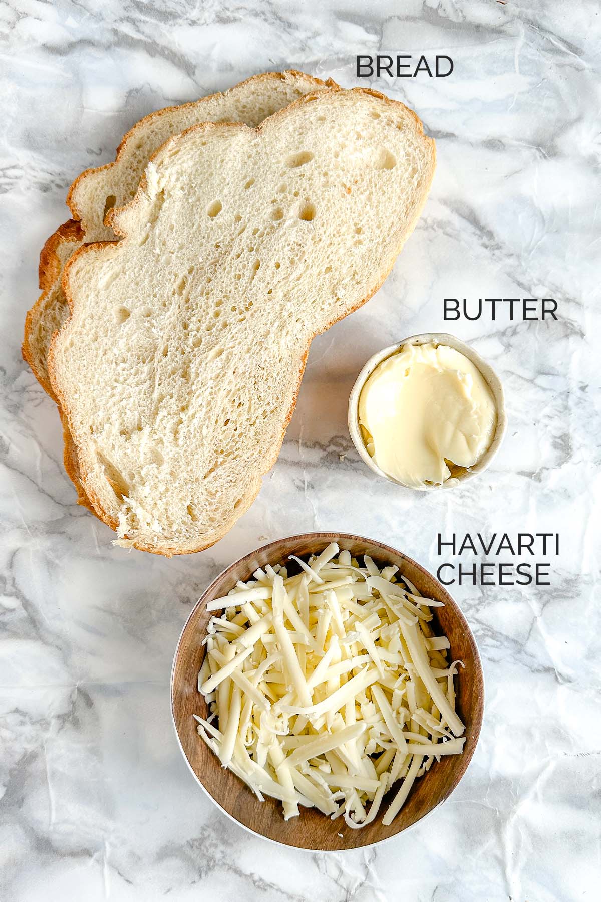 2 slices of bread, a bowl with butter, and a bowl with shredded cheese on a white marble counter.