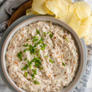 A yogurt dip in a bowl with onions in it and green onions on top beside potato chips.